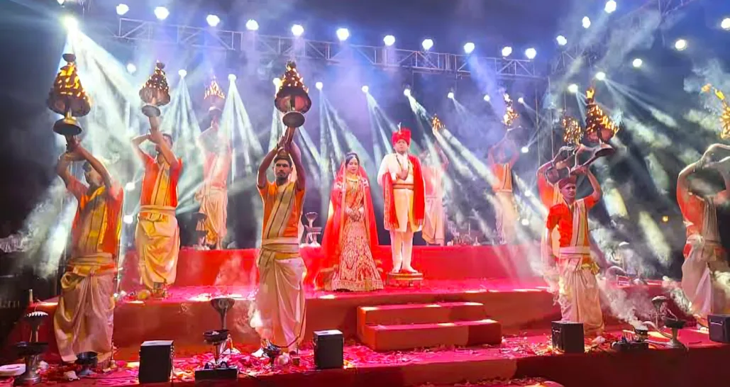 Bride and groom on stage during Ganga Aarti Event with priests holding lit diyas